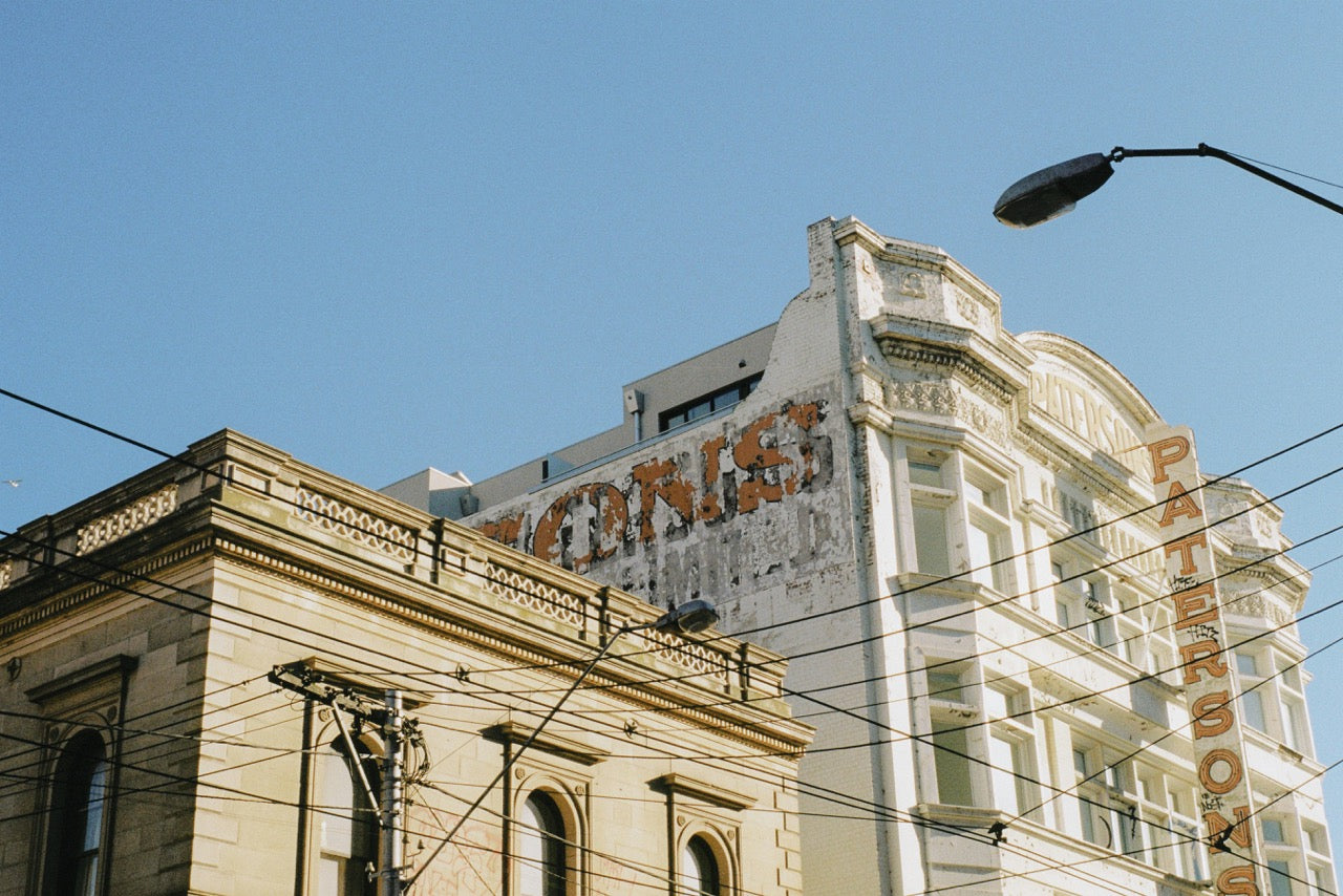 A photograph of historic buildings in Melbourne with overhead tram wires crisscrossing the sky, adding to the city's urban charm under a clear blue sky.