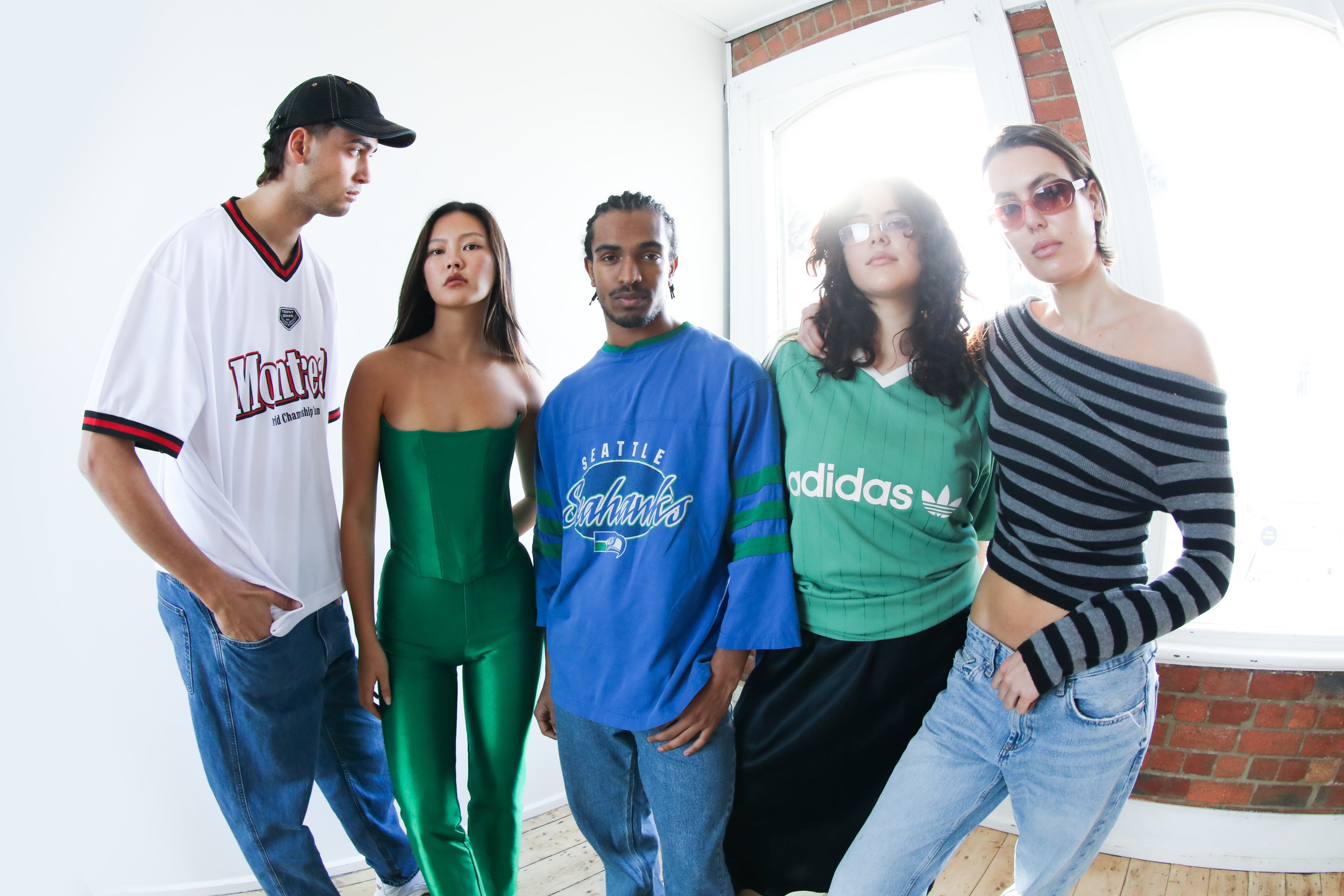 Group of five stylish young adults wearing a mix of vintage and modern secondhand fashion, including streetwear and designer pieces, standing confidently in a naturally lit room with wooden floors and brick walls.