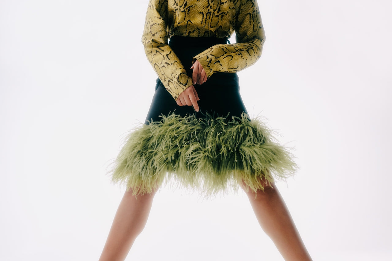 Person wearing a snake-print long-sleeve top paired with a black mini skirt trimmed with green feathers, posing against a white background.