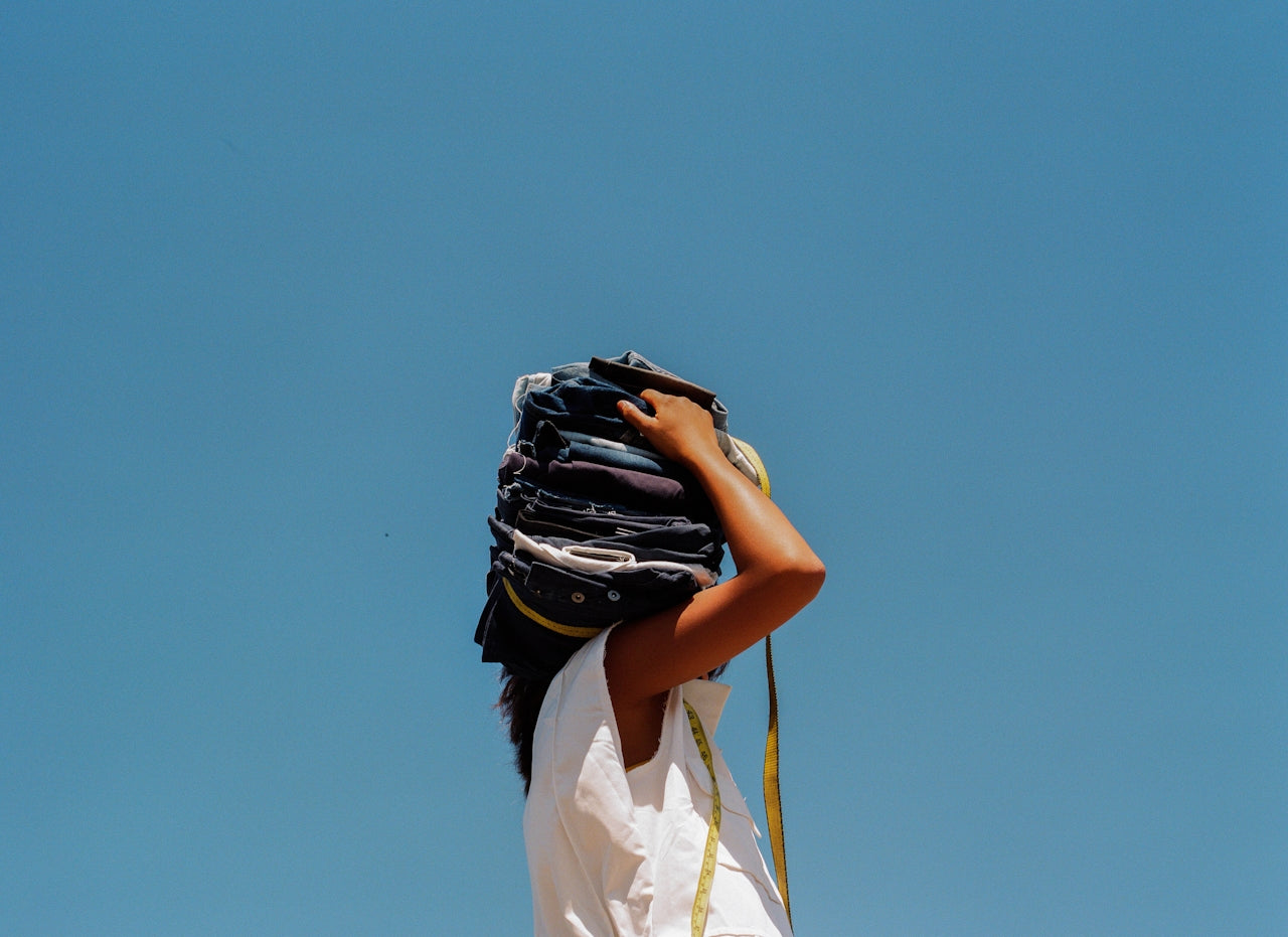 A person wearing a white sleeveless top stands against a clear blue sky, holding a large stack of folded denim jeans balanced on their head, partially covering their face.