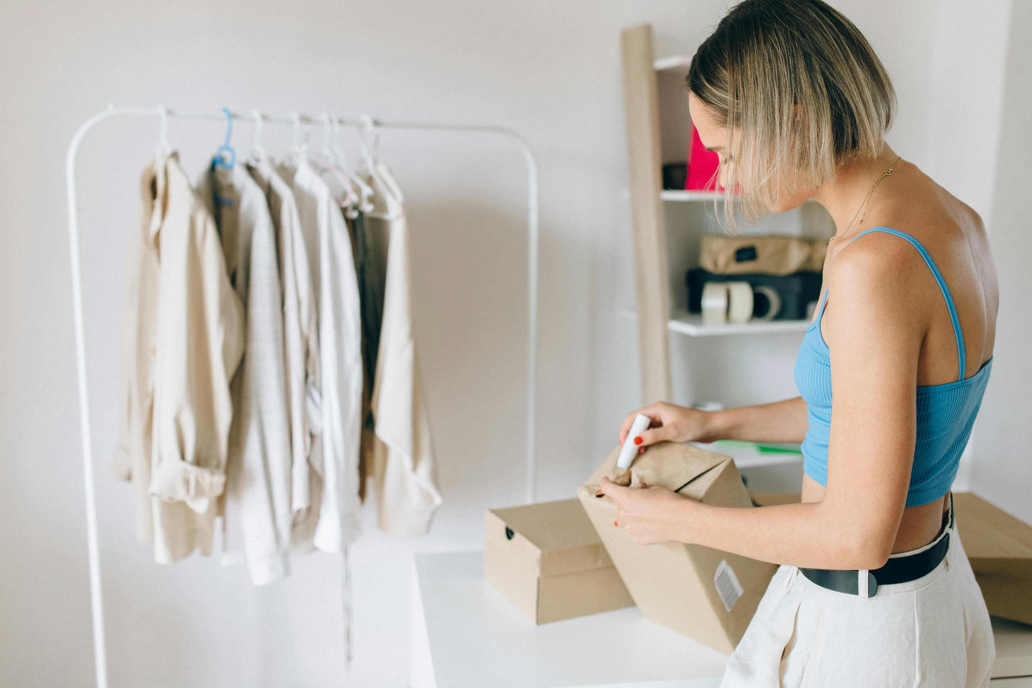Woman in blue tank top and white pants packaging a cardboard box with a clothing rack of neautral-toned garments and shelves of folded clothes in the background.
