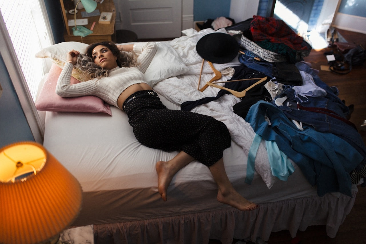 A woman lounges on a bed, surrounded by a pile of clothes, hangers, and accessories.