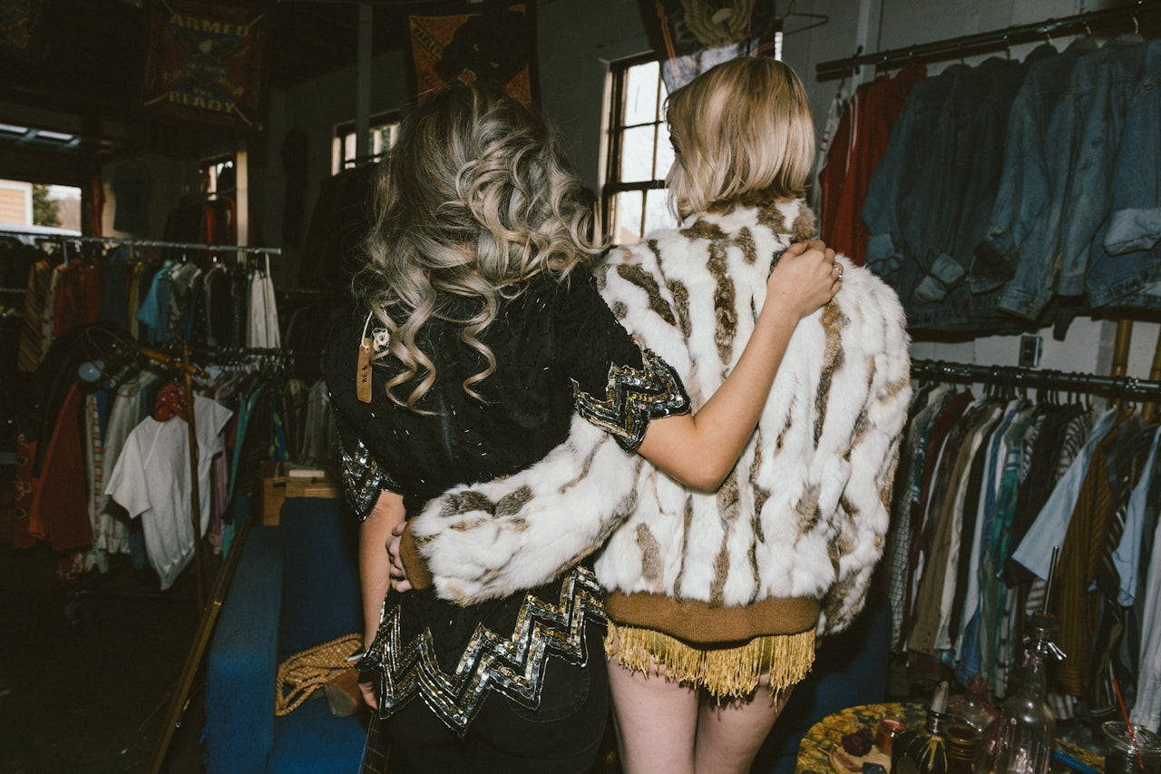 Two stylishly dressed women with their arms around each other browse a vintage clothing store.